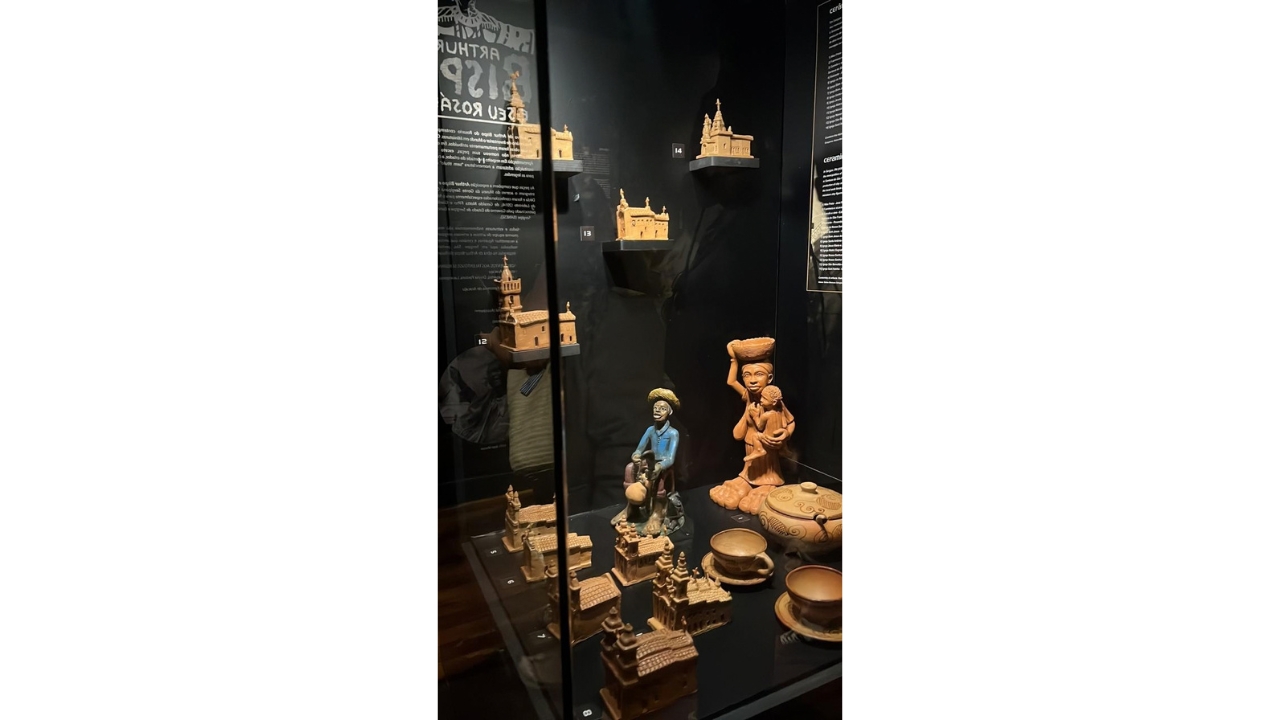 Vitrine de exposição em uma sala escura, de paredes pretas. Dentro dela, há esculturas de figuras humanas, peças de cerâmica e miniaturas arquitetônicas em tons terrosos. O fundo preto contém textos informativos e a numeração das peças expostas.