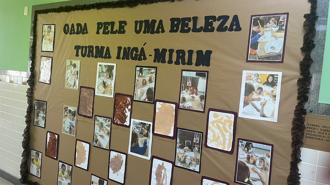 Painel escolar de fundo marrom, com fotografias e produções artísticas infantis. No alto, lê-se, em letras pretas recortadas, “Cada pele uma beleza – Turma Ingá-Mirim”. As imagens, com molduras brancas ou marrons, mostram crianças realizando atividades de pintura, e também pinturas em diferentes tons de marrom. A parede ao fundo é verde com a metade inferior de azulejos brancos.