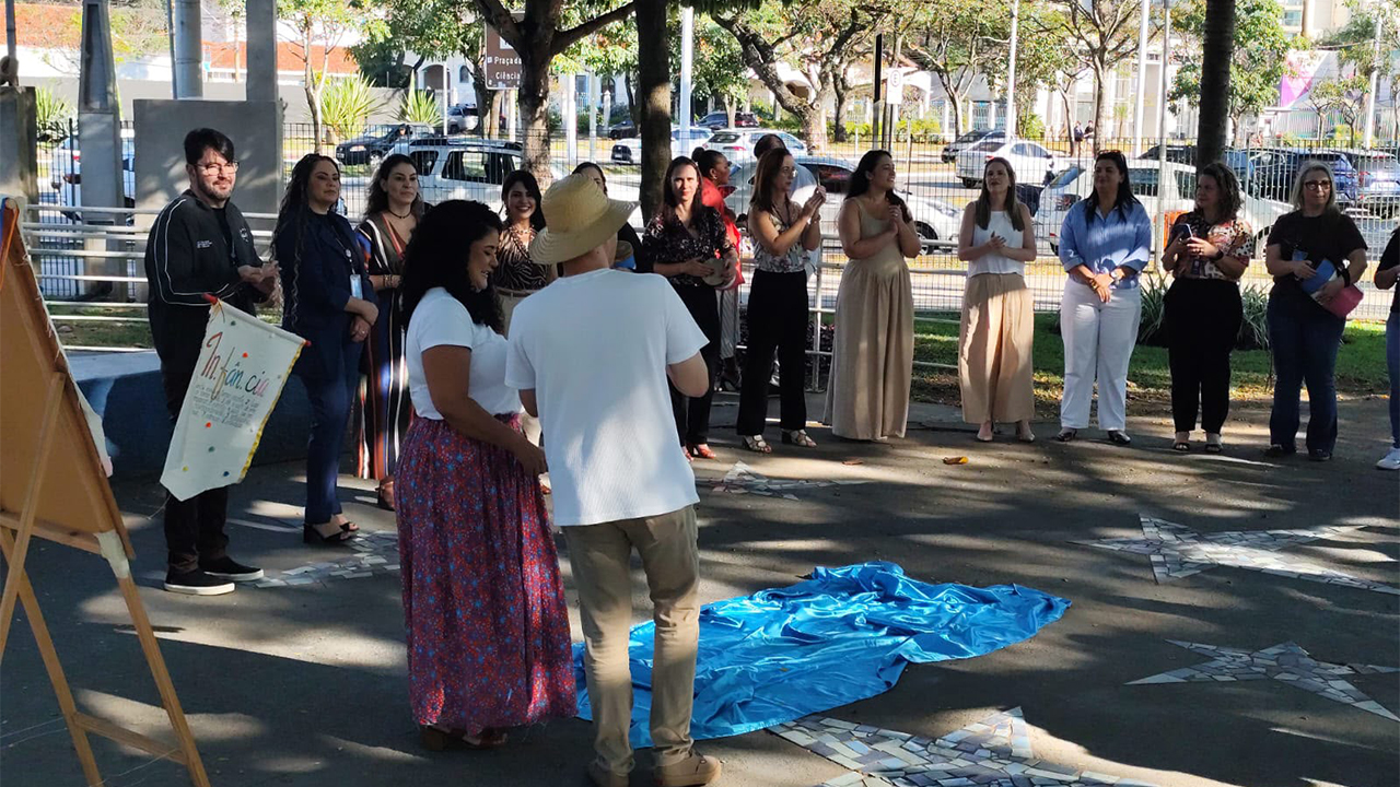 Atividade coletiva ao ar livre, sob árvores, com um grupo multirracial de 13 adultos reunidos em semicírculo. Ao centro, de costas para a câmera, duas pessoas - um homem e uma mulher - conduzem a interação ao lado de um tecido azul estendido no chão de cimento com estrelas de mosaicos. Ao fundo, aparecem uma rua, carros, vegetação e edificações.