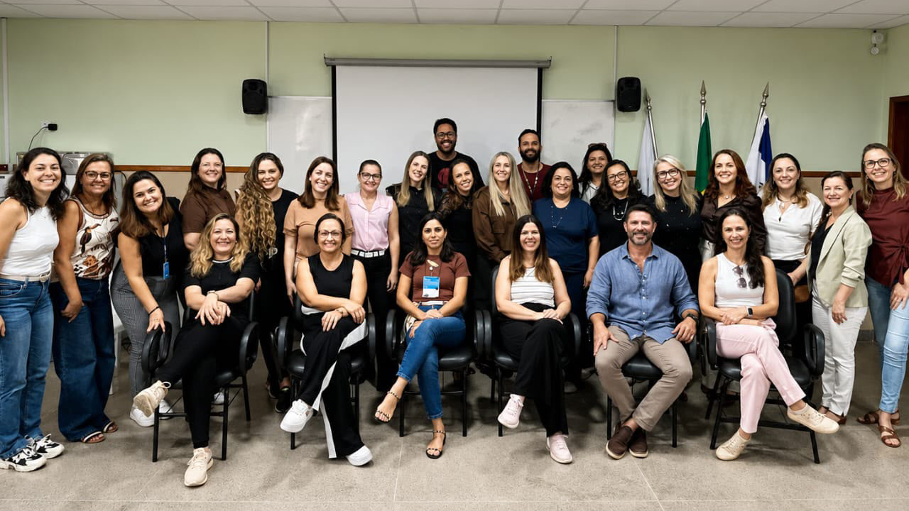 Grupo multirracial de cerca de 26 adultos posa para foto em uma sala de reunião, sendo que seis pessoas estão sentadas à frente e o restante em pé, atrás. O grupo usa roupas casuais em tons variados. Ao fundo, há uma tela de projeção desligada, uma caixa de som preta de cada lado da tela e três bandeiras do lado direito. A parede da sala é verde clara e o chão é cinza. No teto, um projetor está centralizado.