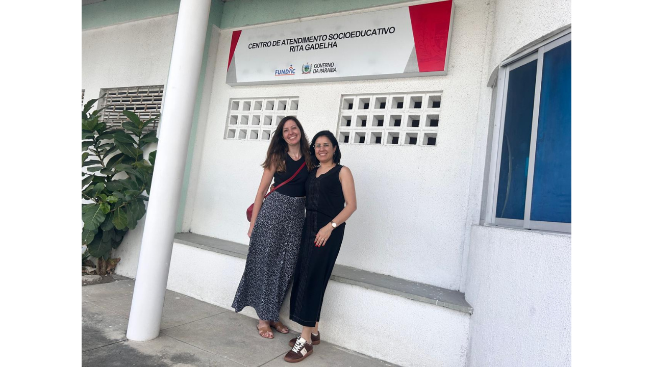 Duas mulheres posam em pé, lado a lado e sorrindo. A da esquerda veste saia longa estampada e blusa preta, e a da direita usa um conjunto preto de calça e blusa. Acima delas, uma placa branca fixada exibe os textos "Centro de Atendimento Socioeducativo Rita Gadelha", com as logomarcas "Fundac" e "Governo da Paraíba". O fundo é composto por uma parede branca texturizada, com elementos vazados de ventilação, um pilar à esquerda e uma janela com fundo azul à direita.