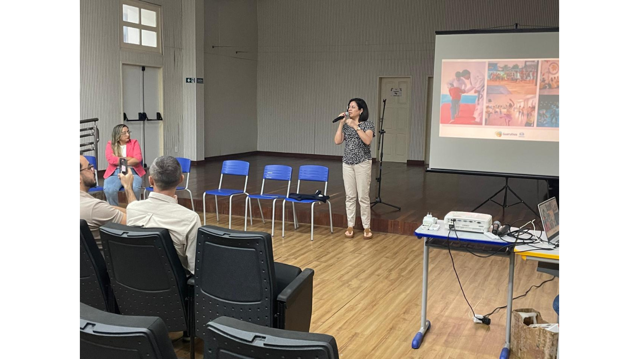 No centro de um auditório, uma mulher de blusa estampada preta e branca e calça clara fala ao microfone, enquanto pessoas sentadas na plateia a observam. À direita, um telão de projeção exibe fotografias coloridas, ao lado de uma mesa com um projetor e notebook. O espaço possui piso de madeira, cadeiras azuis e pretas enfileiradas, portas brancas e paredes claras com textura ripada.