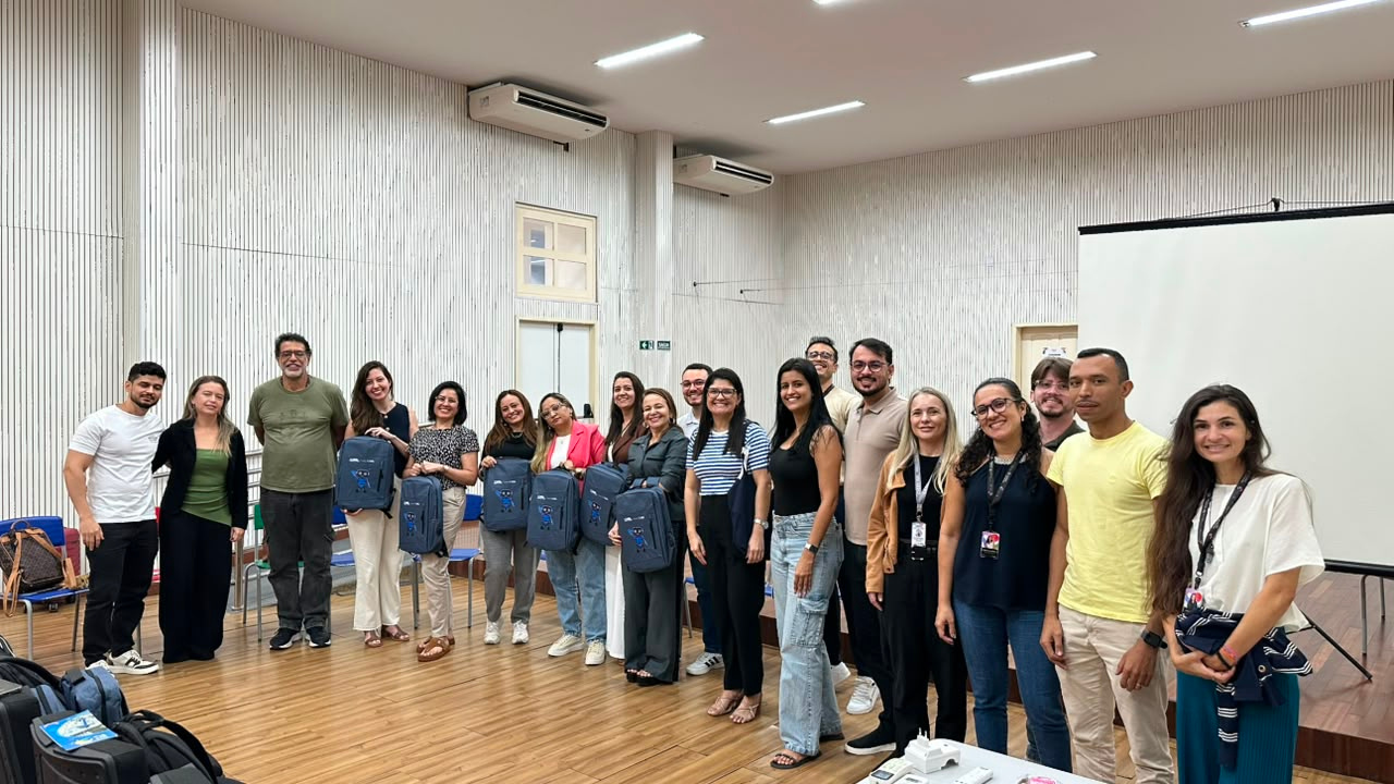Grupo multirracial de cerca de vinte pessoas posa em pé para a foto, em um auditório iluminado, usando roupas casuais variadas. Algumas, no centro do grupo, seguram mochilas azuis idênticas à frente do corpo, com o desenho de um mascote impresso. O espaço conta com piso de madeira, um telão branco à direita e paredes brancas com frisos verticais, além de duas grandes máquinas de ar-condicionado próximas ao teto.