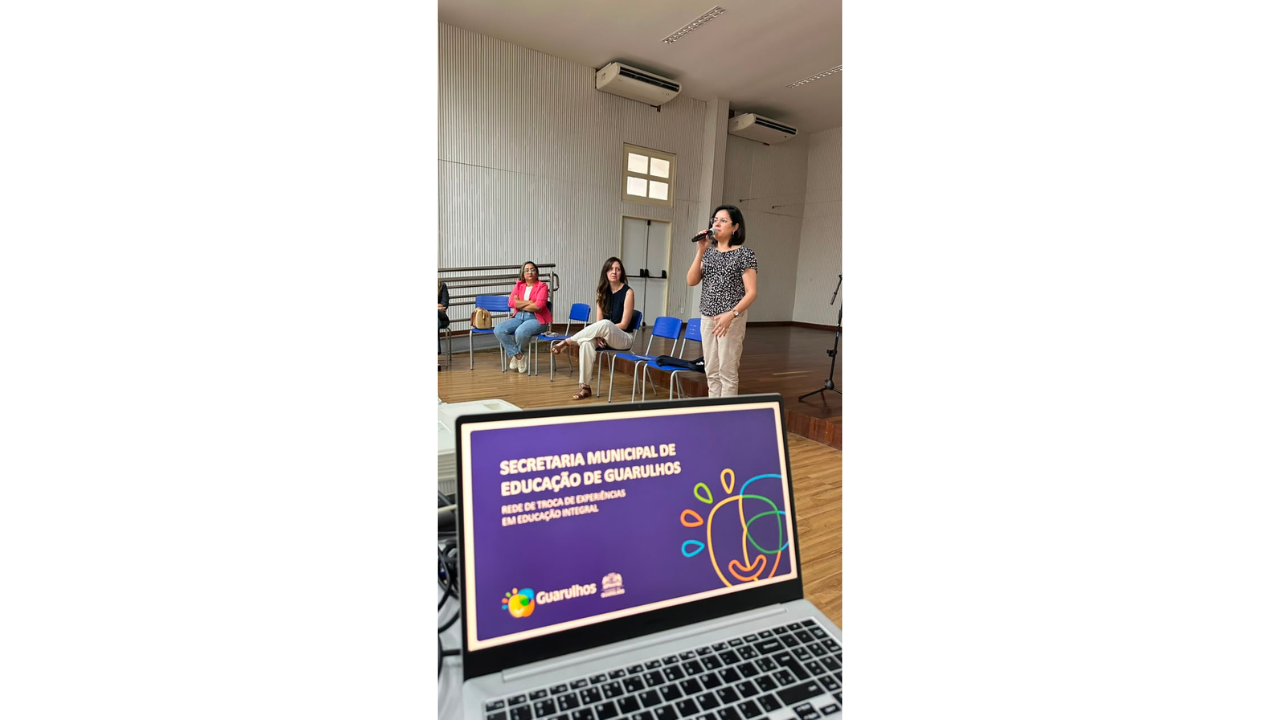 Um notebook exibe uma apresentação com fundo roxo e título "Secretaria Municipal de Educação de Guarulhos", enquanto, atrás dele e ao centro, uma mulher em pé, de blusa estampada, fala ao microfone. Outras duas mulheres, com roupas casuais, estão sentadas ao fundo em cadeiras azuais e a observam. O ambiente possui piso de madeira e parede clara com textura ripada.