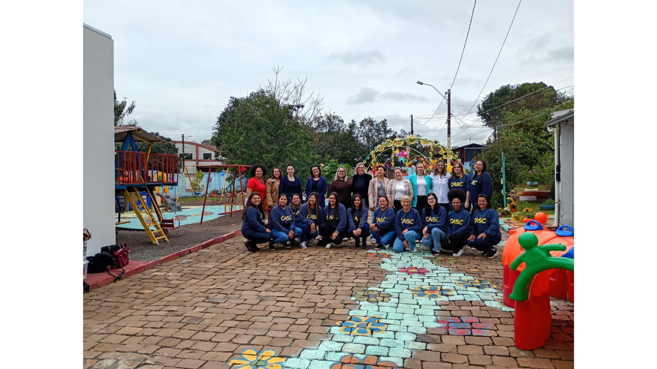 Grupo multirracial de cerca de 23 mulheres posa lado a lado, em pé e agachadas. Várias vestem blusas azul-marinho com a sigla “CASC”em amarelo. O grupo está em um pátio ao ar livre, com um chão de pedras onde está pintado um caminho azul claro com flores coloridas. Ao fundo, há brinquedos, árvores e um arco decorado com flores.