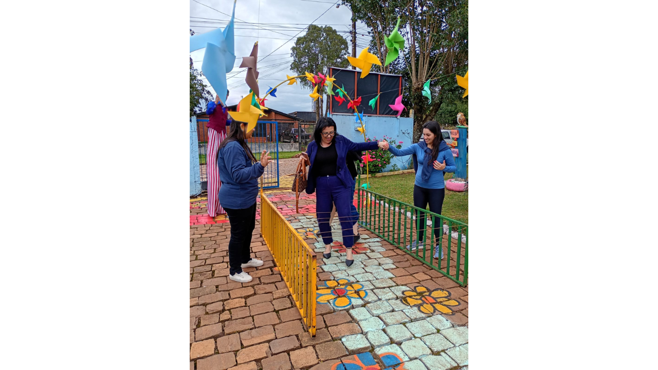 Em uma área externa com um caminho pintado no chão de pedras, três mulheres brincam entre si. A mulher ao centro, vestindo um conjunto azul royal, segura a mão de outra enquanto caminha pelo percurso. Ao redor, há cataventos coloridos, plantas e brinquedos, além de uma figura de perna de pau ao fundo e grades baixas verdes e amarelas.