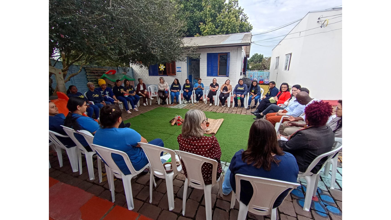 Pátio externo com cerca de vinte e cinco adultos sentados em círculo, em cadeiras brancas. O grupo conversa ao redor de uma área central coberta por grama artificial, onde há objetos e materiais dispostos no chão. Ao fundo, aparecem árvores e uma construção branca com janelas azuis.