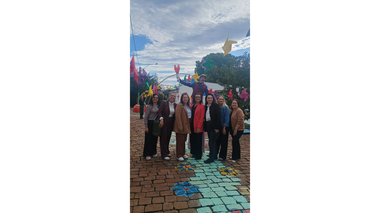 Caminho de pedras decorado com flores pintadas e cataventos coloridos suspensos. Sete mulheres posam lado a lado, usando roupas sociais e casuais em tons variados. Atrás delas, uma pessoa de perna de pau segura objetos de malabarismo.