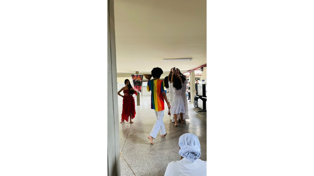 Corredor escolar durante uma apresentação cultural, com grupo multirracial de jovens descalços e usando roupas brancas, vermelhas e uma camiseta multicolorida. Alguns caminham, enquanto outros erguem objetos de barro acima da cabeça. Ao fundo, há cartazes, colunas e pessoas observando a atividade.