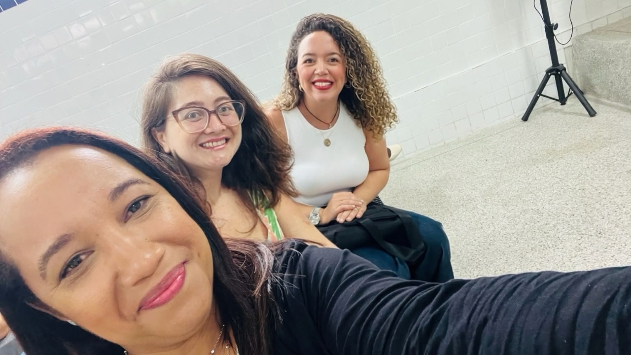 Grupo multiracial composto por três mulheres em um ambiente interno. Elas estão sentadas próximas umas das outras, posando para uma selfie e sorrindo para a câmera. Uma veste roupa preta, outra usa óculos e roupa estampada, e a terceira veste blusa branca. Ao fundo, há parede clara, piso cinza e um tripé.