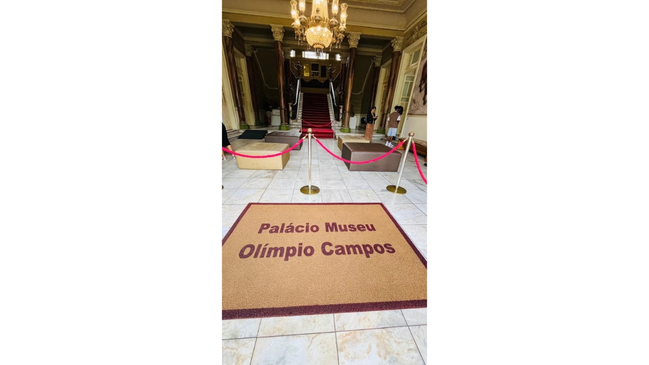 Hall interno de um edifício histórico, com escadaria central coberta por tapete vermelho e delimitada por cordões. Em primeiro plano, um tapete traz a inscrição “Palácio Museu Olímpio Campos”. Ao fundo, destacam-se colunas, lustre e portas.