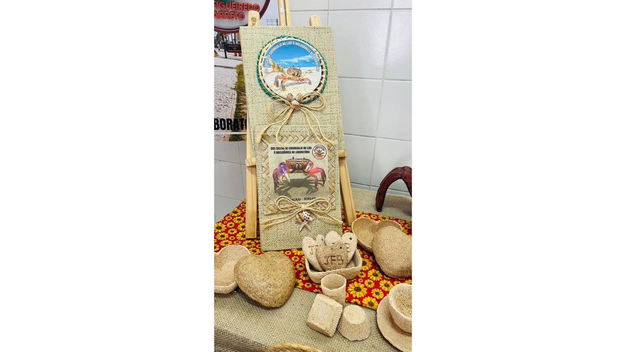Exposição pedagógica sobre o reaproveitamento de cascas de caranguejo, organizada sobre uma mesa forrada com toalha trançada e uma toalha estampada, com fundo vermelho e flores de girassol. Um cavalete exibe o texto “Das cascas de caranguejo no lixo à biocerâmica no laboratório”, acompanhado de imagens de caranguejos. Ao redor, há recipientes e objetos de tonalidade bege produzidos com o material.