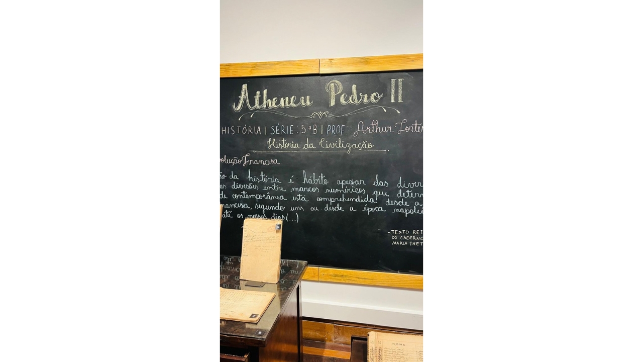 Sala de exposição histórica com uma lousa preta de giz com molduras de madeira e carteiras antigas. No quadro, lê-se “Atheneu Pedro II”, além de anotações feitas a giz branco. Em primeiro plano, há cadernos e documentos antigos sobre mesas de madeira.
