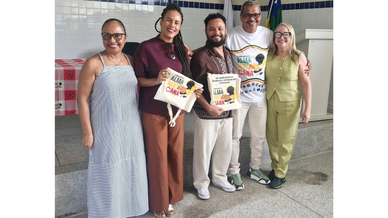 Grupo multirracial de cinco adultos posa lado a lado em uma sala. Duas pessoas seguram sacolas beges (ecobags) com a inscrição “Projeto Alma Africana”, e um homem veste uma camiseta branca com o mesmo nome. Ao fundo, há bandeiras, uma mesa decorada com toalha xadrez rosa e branca e uma parede revestida com azulejos brancos e azuis.