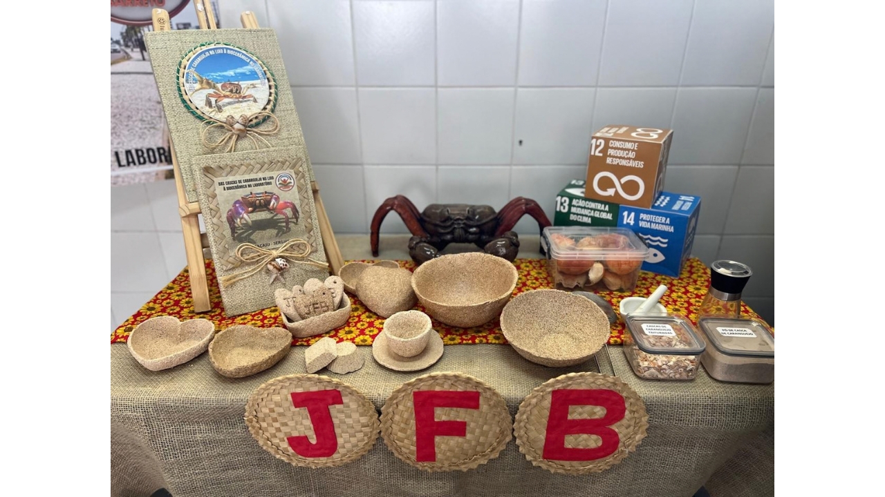 Exposição pedagógica sobre o reaproveitamento de cascas de caranguejo, organizada sobre uma mesa, forrada com uma toalha trançada e outra com fundo vermelho e estampa de girassóis. Sobre a mesa, há recipientes e objetos beges produzidos com o material, além de um cavalete e três caixas marrom, verde e azul referentes aos ODS 12, 13 e 14: Consumo e produção responsáveis, Ação contra a mudança global do clima e Proteção a vida marinha, respeticamente. Na frente da mesa, três peças circulares, aparentemente de palha trançada, trazem as letras “J”, “F” e “B” em vermelho.