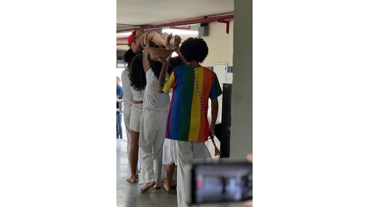 Corredor escolar durante uma apresentação cultural, com participantes descalços usando roupas brancas e uma camiseta listrada multicolorida. Um grupo ergue uma participante e um recipiente de barro acima da cabeça. Em primeiro plano, um celular fotografa a ação.