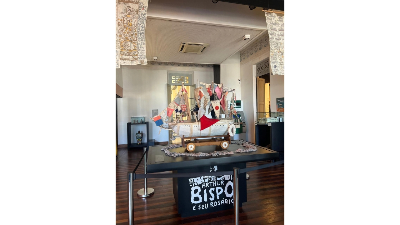 Ambiente de exposição com uma embarcação artística feita de tecidos, cordas, bandeiras e estruturas de madeira. A obra está sobre uma mesa preta, apoiada em uma plataforma também preta, cercada por barreiras de proteção, com a inscrição na cor branca: “Arthur Bispo e seu Rosário” na parte frontal. Ao fundo, há outras peças expostas, portas e painéis decorativos. O teto do ambiente é branco e o chão de madeira escura.