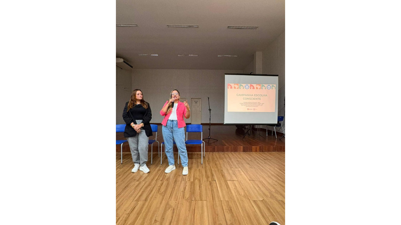 Em uma sala de apresentação, duas mulheres estão em pé diante do público. Uma delas, de blazer rosa, fala ao microfone; a outra veste blazer preto e observa. Ao lado, uma tela exibe o título “Campanha Escolha Consciente”. A sala tem piso de madeira clara, teto branco e paredes brancas. Ao fundo, há cadeiras azuis e um palco de madeira.