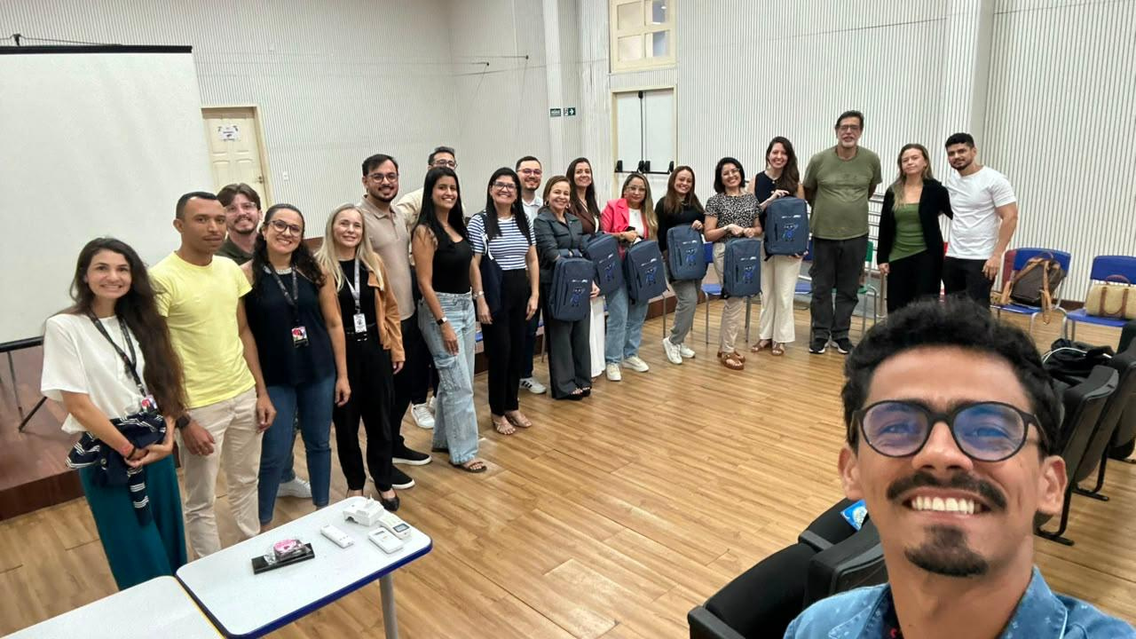 Grupo multirracial de 20 adultos reunidos para uma fotografia. Em primeiro plano, um homem faz uma selfie enquanto o grupo posa ao fundo. Algumas pessoas seguram uma mochilha azul, igual. O ambiente tem piso de madeira, cadeiras azuis, mesas e paredes claras. Ao fundo, há uma tela branca de projeção desligada.