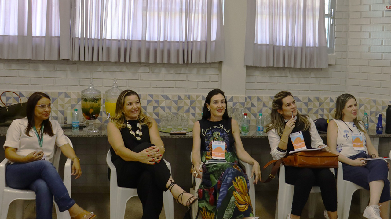 Ambiente interno com cinco mulheres sentadas lado a lado em cadeiras brancas organizadas em semicírculo, acompanhando uma atividade. Elas usam roupas casuais, algumas com crachás, e mantêm a atenção voltada para fora do enquadramento. Ao fundo, há uma bancada com recipientes de vidro de bebidas, copos, garrafas de água e janelas com cortinas claras. A parede é de tijolos brancos aparentes e azulejos com triângulos azuis e beges.