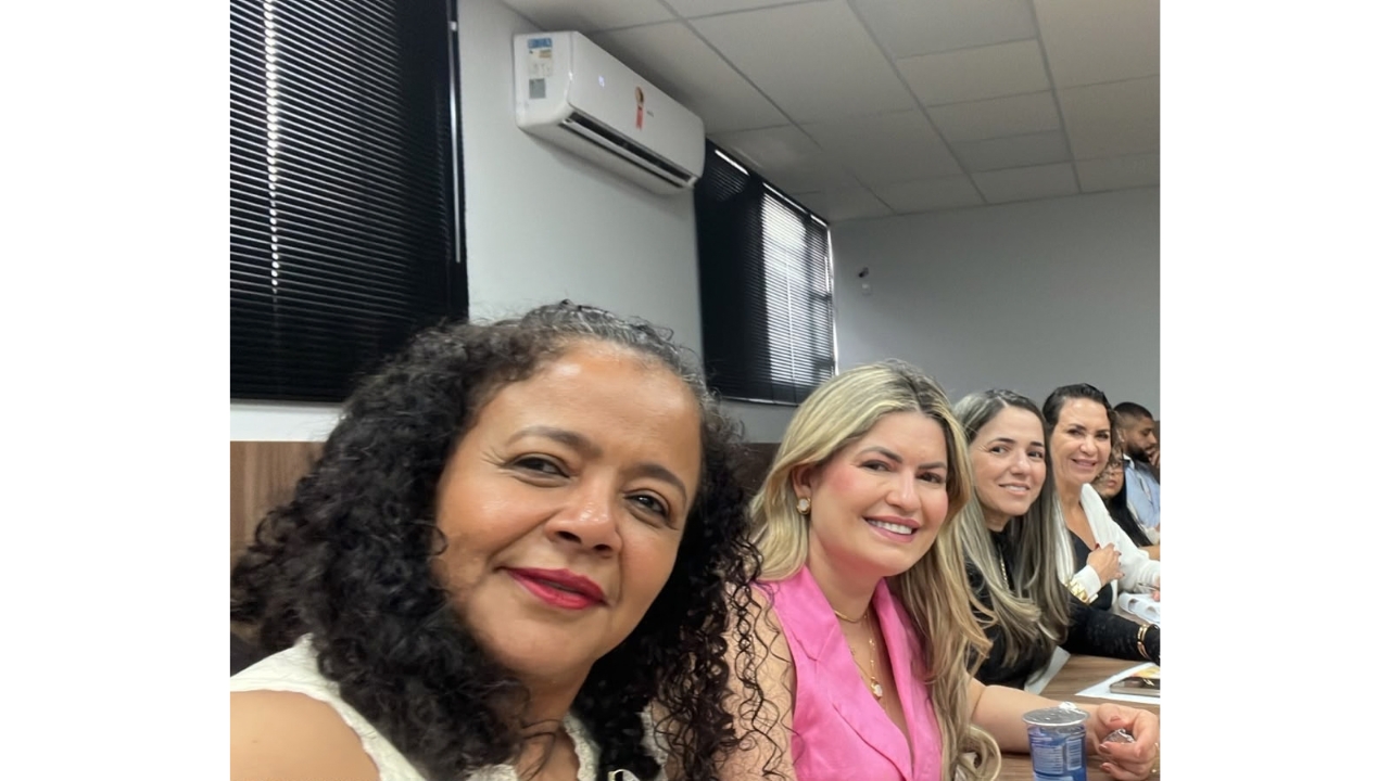 Sala de reunião com várias pessoas sentadas lado a lado à mesa. Em primeiro plano, um grupo multirracial de quatro mulheres sorri para a câmera. Ao fundo, há outras pessoas sentadas. O ambiente conta com janelas com persianas pretas e um aparelho de ar-condicionado.