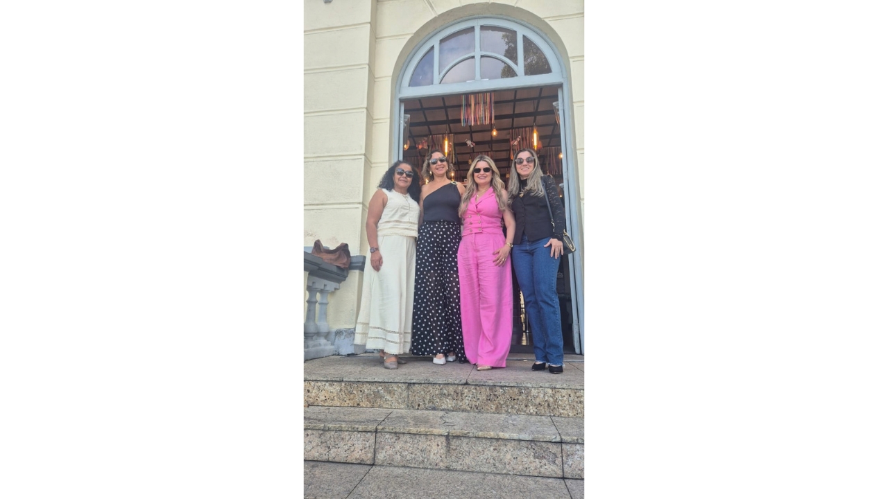 Grupo multirracial de quatro mulheres posa em pé, lado a lado, em frente a uma porta de entrada dupla no formato de arco. Elas usam roupas casuais e as quatro estão de óculos escuros. Ao fundo, vê-se o interior do prédio amarelo claro com elementos decorativos pendentes. À frente delas, dois degraus da escada de acesso.