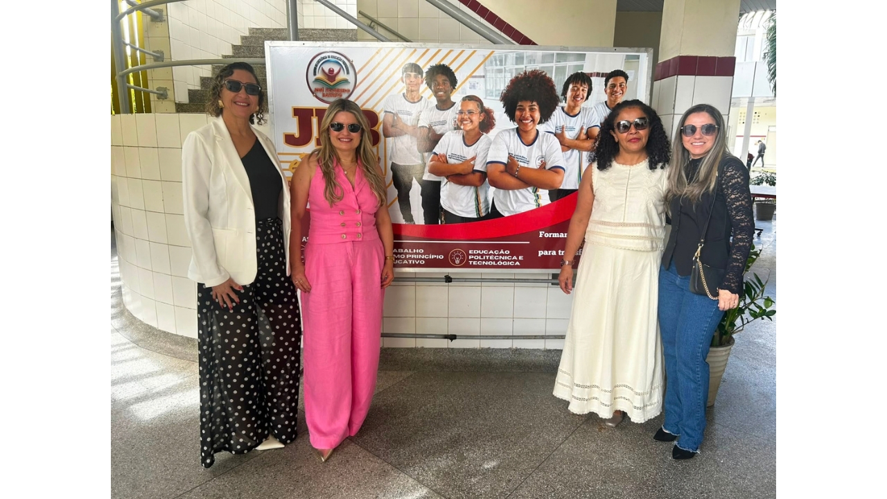 Grupo multirracial de quatro mulheres posa ao lado de um painel informativo em área interna de uma instituição. Elas usam óculos escuros e roupas sociais e casuais, incluindo peças brancas, pretas e rosa. O painel exibe imagens de estudantes e textos sobre educação; ao fundo, há escadas, paredes revestidas de azulejos claros e iluminação natural.