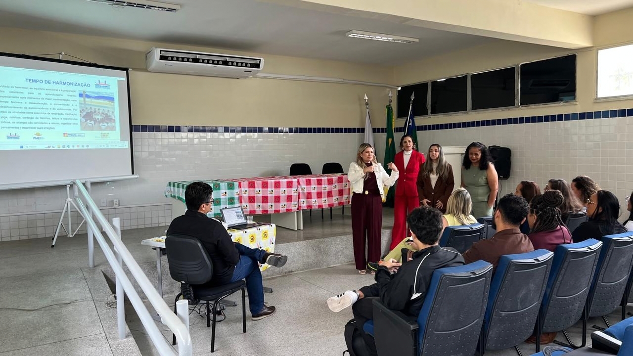 Sala de apresentação com público sentado em cadeiras pretas com estofado azul voltadas para a frente. Um grupo multirracial de quatro mulheres está em pé à frente, enquanto uma delas gesticula durante a fala. À esquerda, há uma tela com o título "Tempo de Harmonização"; à direita há uma mesa com toalhas xadrez rosa e verde, além de três bandeiras. As paredes da sala são amarelas e revestidas com azulejos brancos e azul marinho.