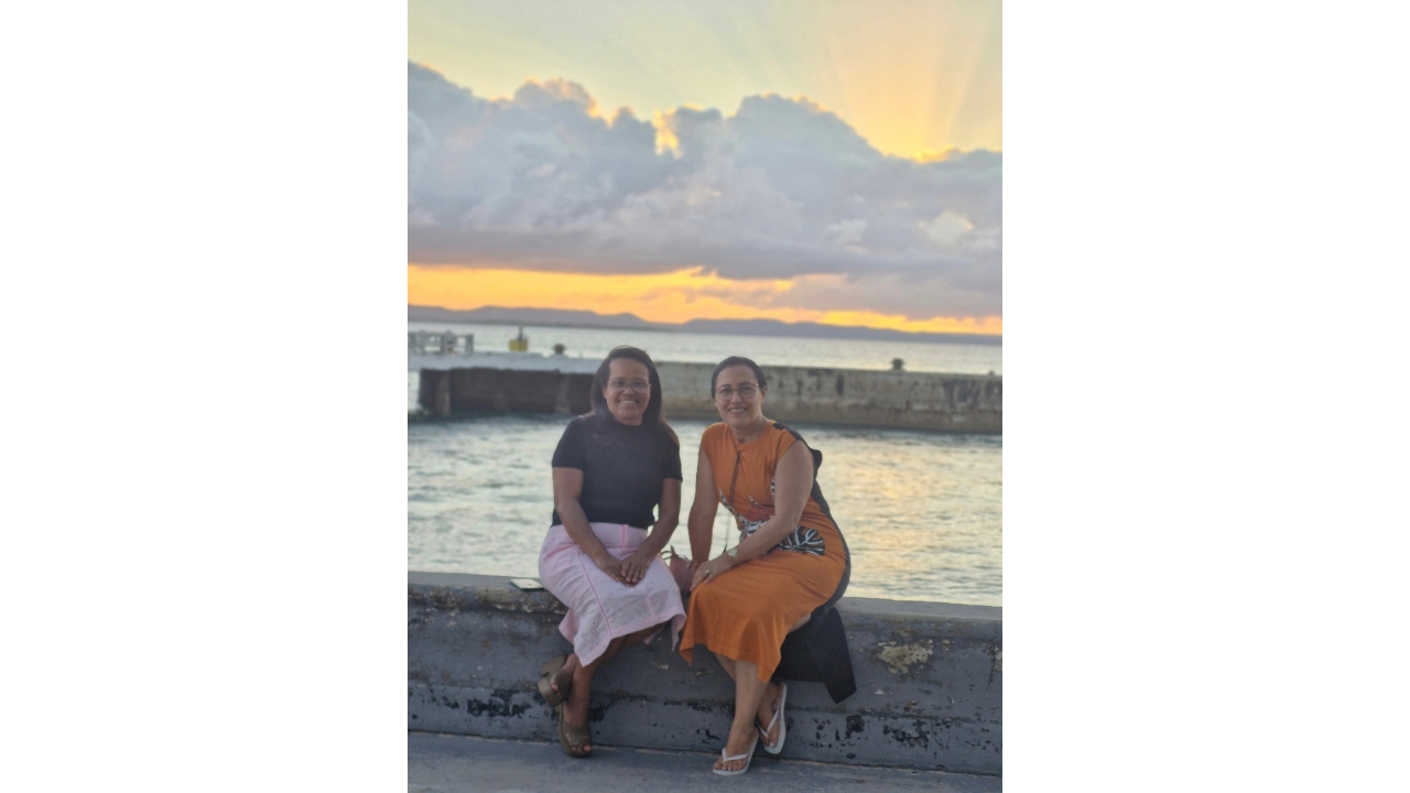 Área externa à beira-mar durante o pôr do sol. Duas mulheres, sentadas lado a lado em uma mureta de pedra, sorriem para a câmera; uma veste blusa preta e saia rosa, e a outra, vestido laranja. Ao fundo, aparecem o mar, um muro e o céu alaranjado.