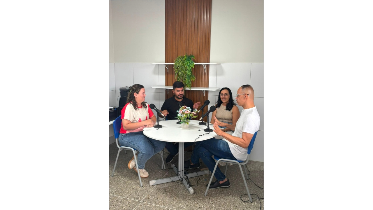 Um grupo multirracial de quatro adultos está sentado ao redor de uma mesa redonda branca, conversando e gesticulando diante de microfones. No centro da mesa, há um vaso bege com flores. Ao fundo, há um painel de madeira, duas prateleiras brancas e uma planta.