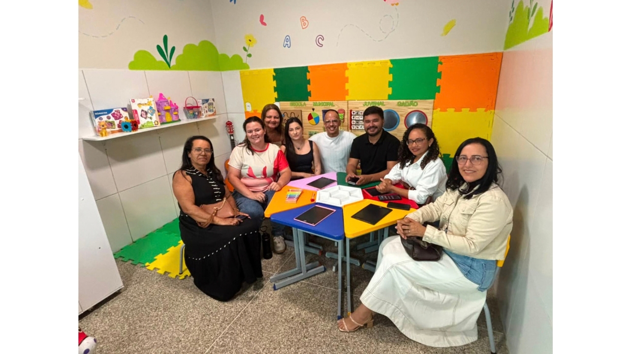 Sala com mobiliário infantil e recursos pedagógicos coloridos. Um grupo multirracial de oito adultos posa ao redor de pequenas mesas triangulares coloridas, sobre as quais há tablets. Ao fundo, aparecem painéis sensoriais, placas de EVA laranjas, amarelas e verdes e brinquedos. No canto esquerdo, uma prateleira branca serve como suporte para livros e brinquedos.