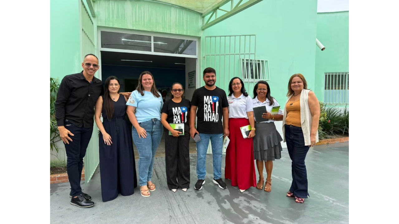 Grupo multirracial de oito adultos posa lado a lado em frente a uma construção verde claro. Algumas pessoas seguram cadernos ou materiais impressos. Duas vestem camisetas pretas iguais com a palavra “Maranhão”. Ao fundo, há um portão, janelas gradeadas e vegetação.