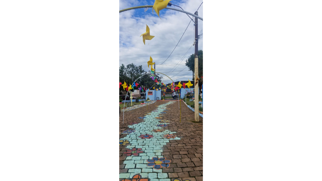 Ao ar livre, um caminho de tijolos vermelhos possui a pintura de uma trilha verde-clara no chão, contornada por flores coloridas, sobre a qual arcos metálicos sustentam diversos cata-ventos de papel colorido. O cenário ao fundo inclui árvores, postes e um prédio azul.