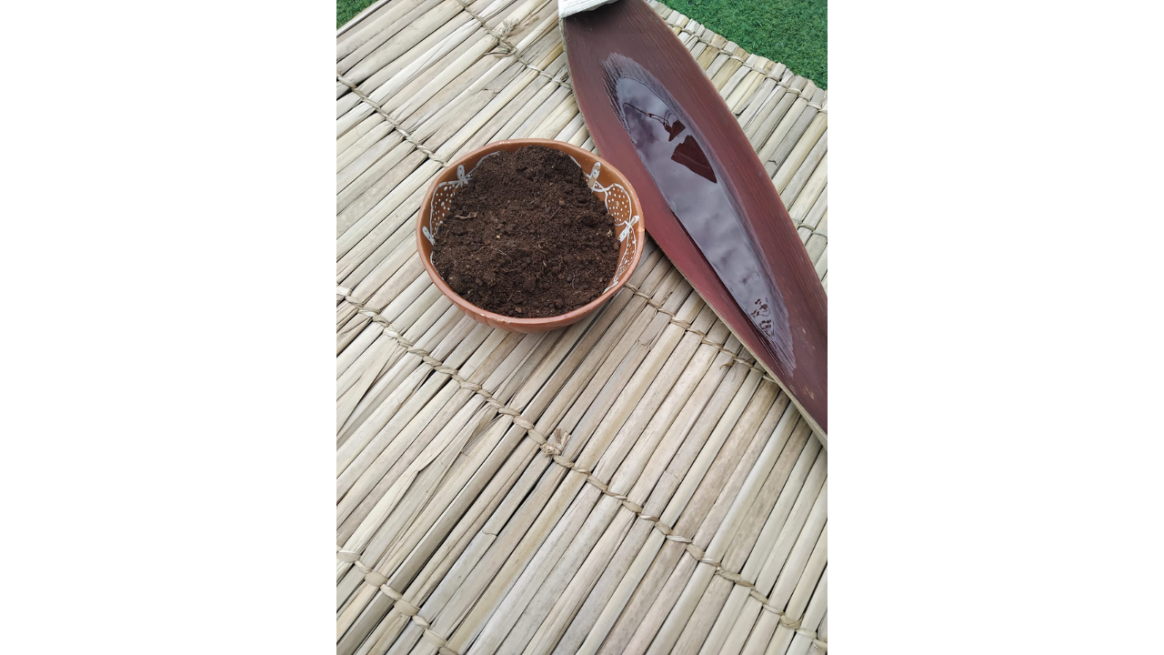 Acima de uma esteira de palha trançada, há uma pequena tigela de cerâmica marrom contendo terra escura. Ao lado, há uma longa folha seca avermelhada, que acumula um pouco de água em seu interior.
