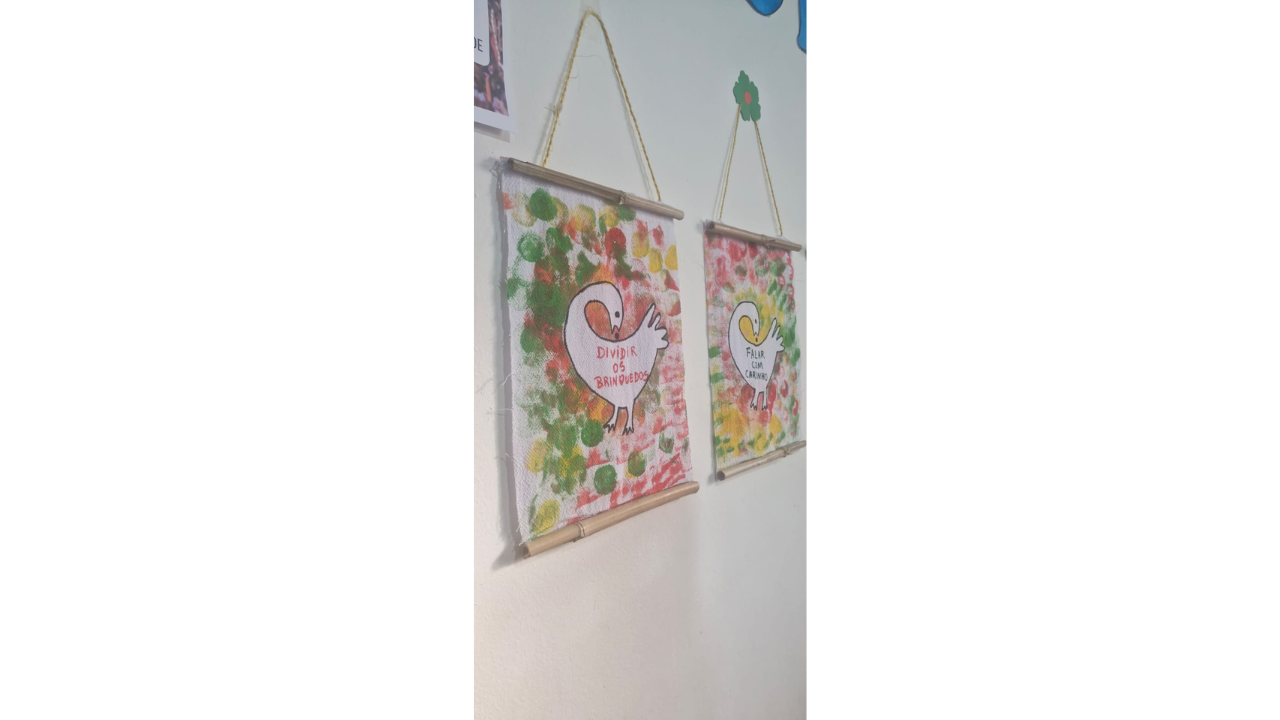 Dois pequenos estandartes artesanais estão pendurados em uma parede branca. Os tecidos possuem hastes de bambu nas extremidades e cordões de fixação. A ilustração consiste em um fundo colorido e uma ave branca ao centro, com uma mensagem em cada estandarte: "Dividir os brinquedos" e "Falar com carinho".