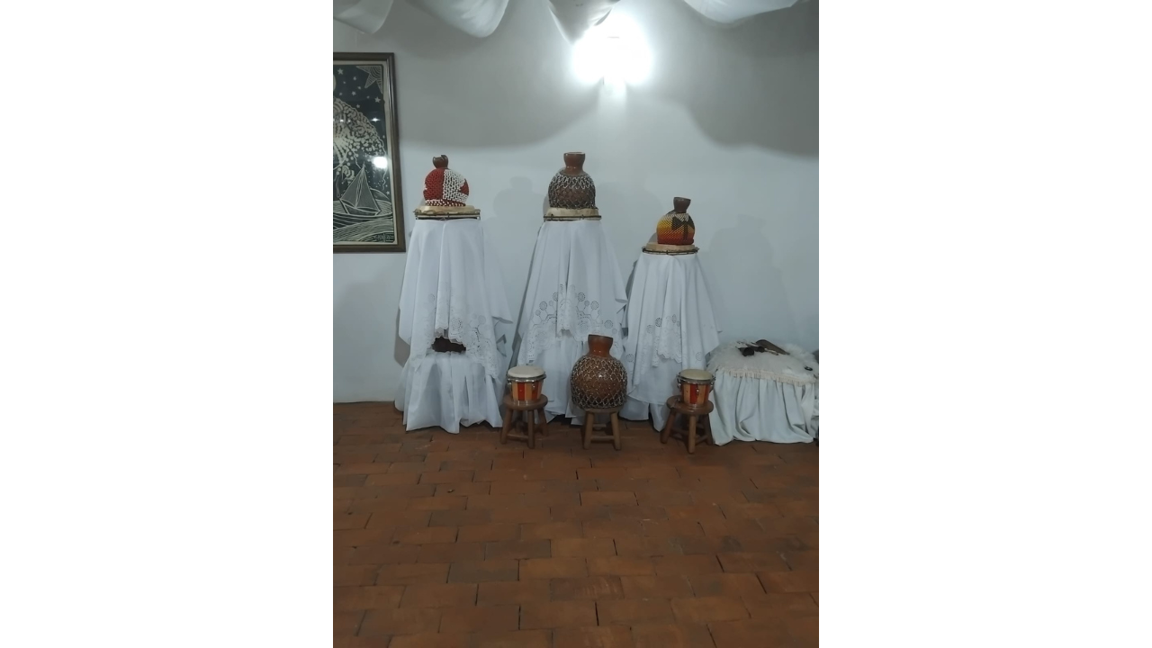 Exposição de instrumentos em uma sala com piso de blocos marrons e paredes brancas. Ao centro, três suportes altos, cobertos com panos brancos rendados, sustentam três grandes xequerês (instrumento musical de percussão). À frente, dois pequenos tambores e outro xequerê repousam em banquinhos de madeira.