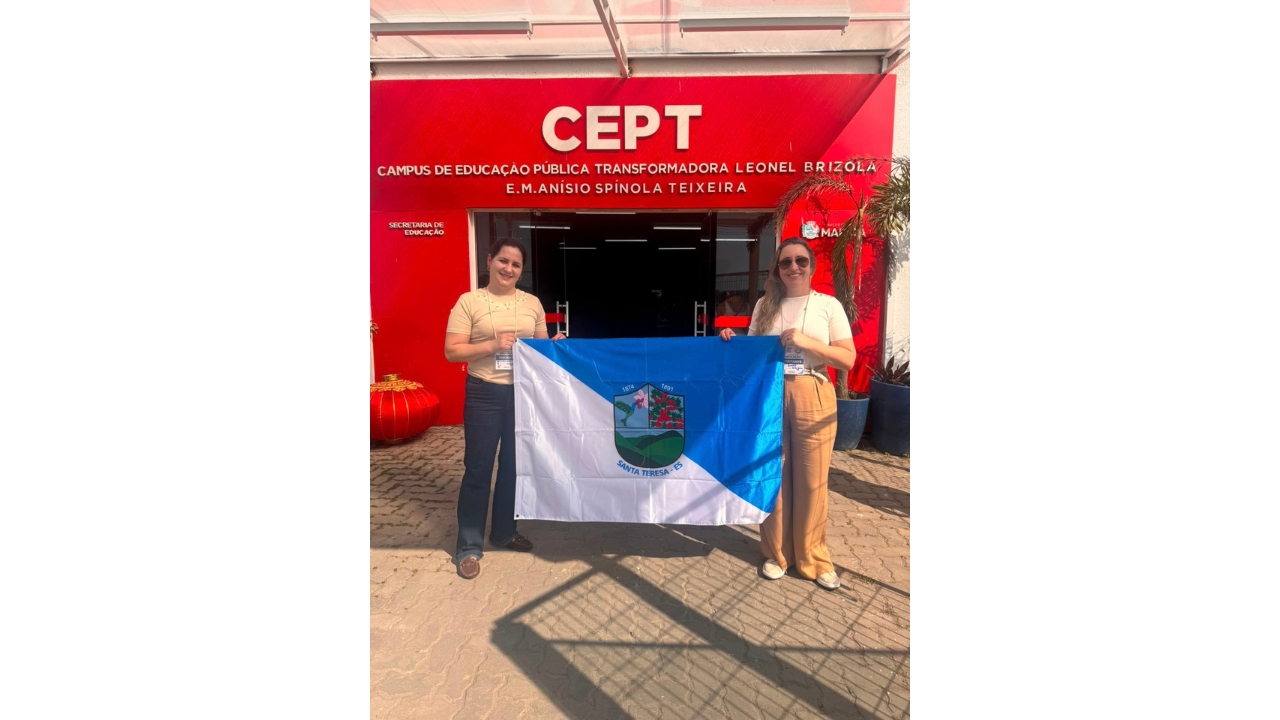 Duas mulheres, usando crachás e roupas casuais, posam segurando as pontas da bandeira de Santa Teresa (ES), dividida diagonalmente entre as cores branca e azul, com um brasão no centro. Ao fundo, a fachada vermelha de um prédio exibe o letreiro branco “CEPT – Campus de Educação Pública Transformadora Leonel Brizola – E.M. Anísio Spínola Teixeira”.