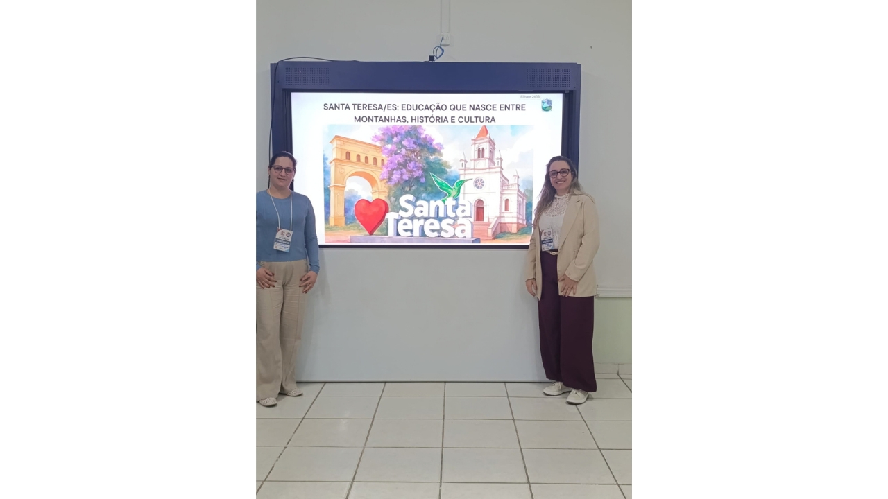 Uma dupla de mulheres de óculos e crachás posam cada uma de um lado de uma tela digital. A tela exibe o texto “Santa Teresa/ES: Educação que nasce entre montanhas, história e cultura”, acompanhado de ilustrações de pontos locais. Ao fundo, há uma parede clara e o piso é de cerâmica clara.