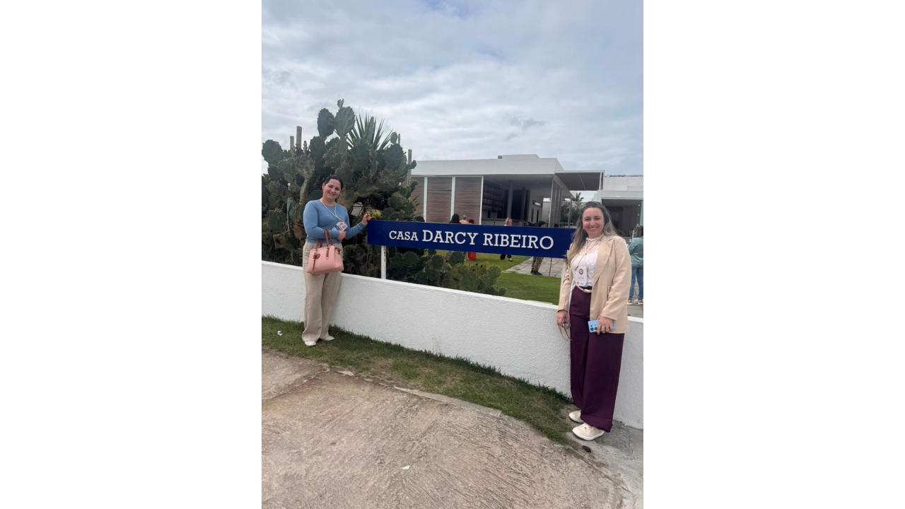 Duas mulheres usando roupas casuais posam ao lado de uma placa azul com a inscrição branca “Casa Darcy Ribeiro”, posicionada na grama acima de uma mureta branca. Ao fundo, há cactos, gramado, uma construção baixa na cor branca e outras pessoas.