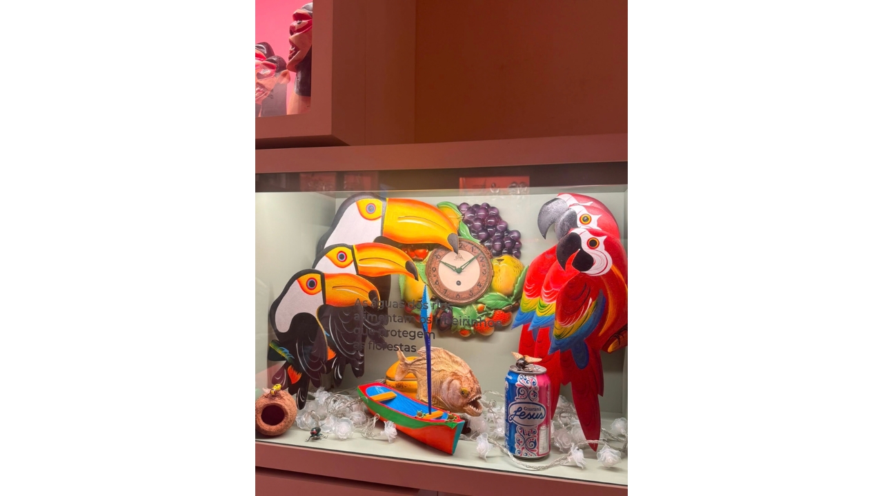 Vitrine de exposição com objetos coloridos relacionados à fauna e à cultura amazônica. Destacam-se três figuras de tucanos e três araras, posicionadas em tamanho crescente, um peixe, uma pequena embarcação colorida, um relógio e uma lata de guaraná Jesus. Os objetos estão organizados atrás de uma proteção de vidro.