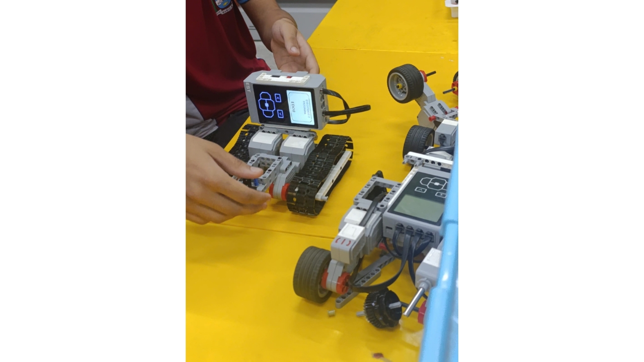 Mesa amarela com equipamentos de robótica educacional. Em primeiro plano, uma pessoa vestindo camiseta na cor bordô manuseia um robô com esteiras e módulo eletrônico com tela, enquanto outro robô com rodas está ao lado. O cenário destaca peças, cabos e componentes de montagem.