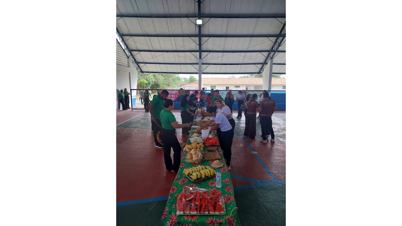 Em uma quadra poliesportiva coberta, adultos estão reunidos em torno de uma mesa comprida. Sobre a mesa há frutas cortadas, pães, bebidas e outros alimentos, enquanto algumas pessoas se servem. Ao fundo, aparecem outros participantes, grades, árvores e edificações. O teto é inclinado e com forro claro. O piso é pintado de verde e vermelho, com linhas azuis para delimitar os campos de futebol e basquete.