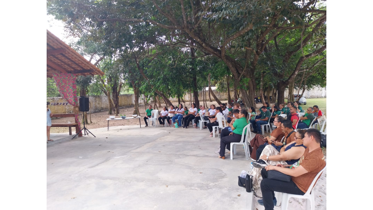 Área externa arborizada com cerca de trinta adultos sentados em cadeiras brancas organizadas em semicírculo, acompanhando uma atividade. O grupo multirracial usa roupas casuais, como calças e camisetas, e está voltado para uma área de apresentação. O espaço tem piso de concreto na cor cinza claro. Uma estrutura à frente tem o teto inclinado, uma cortina com estampa de flores e uma caixa de som preta. Ao fundo, é possível ver um muro cinza e diversas árvores.