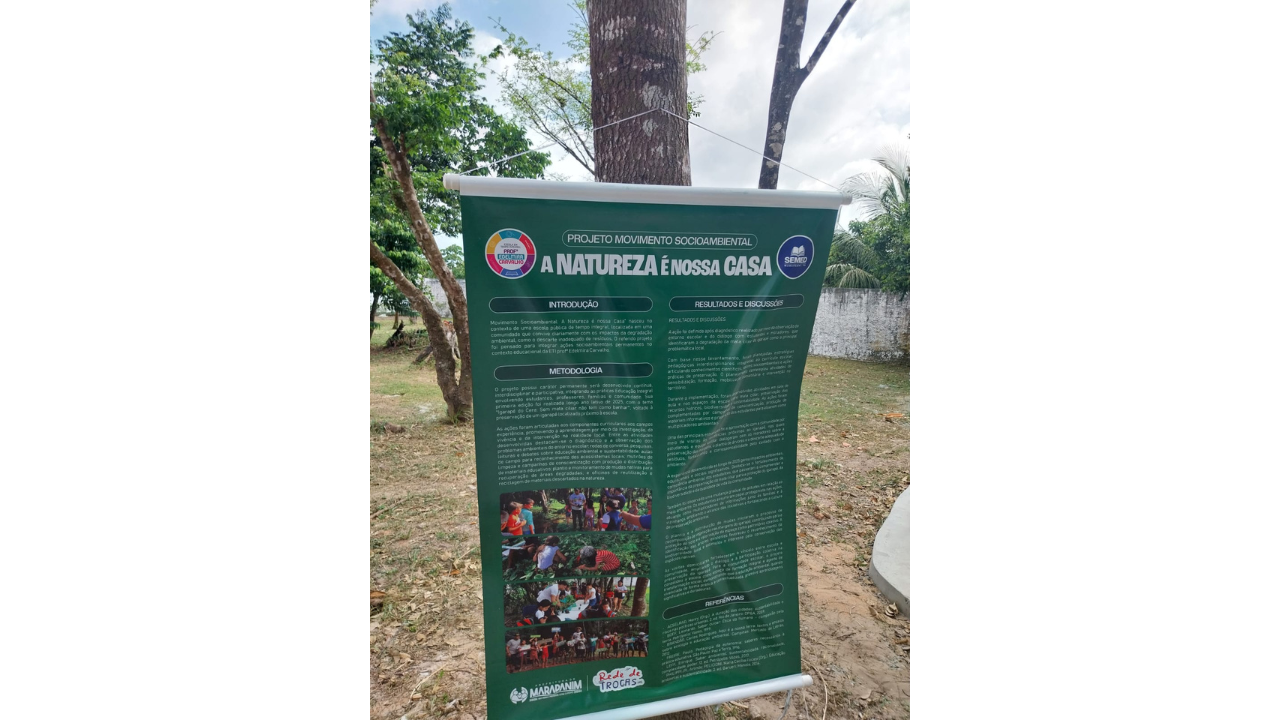 Banner informativo verde pendurado em uma árvore. O material tem o título “Projeto Movimento Socioambiental – A Natureza é Nossa Casa” e é dividido em textos como introdução, metodologia, resultados e referências, além de fotografias de atividades. Ao fundo, há árvores, solo de grama e terra batida e um muro cinza.