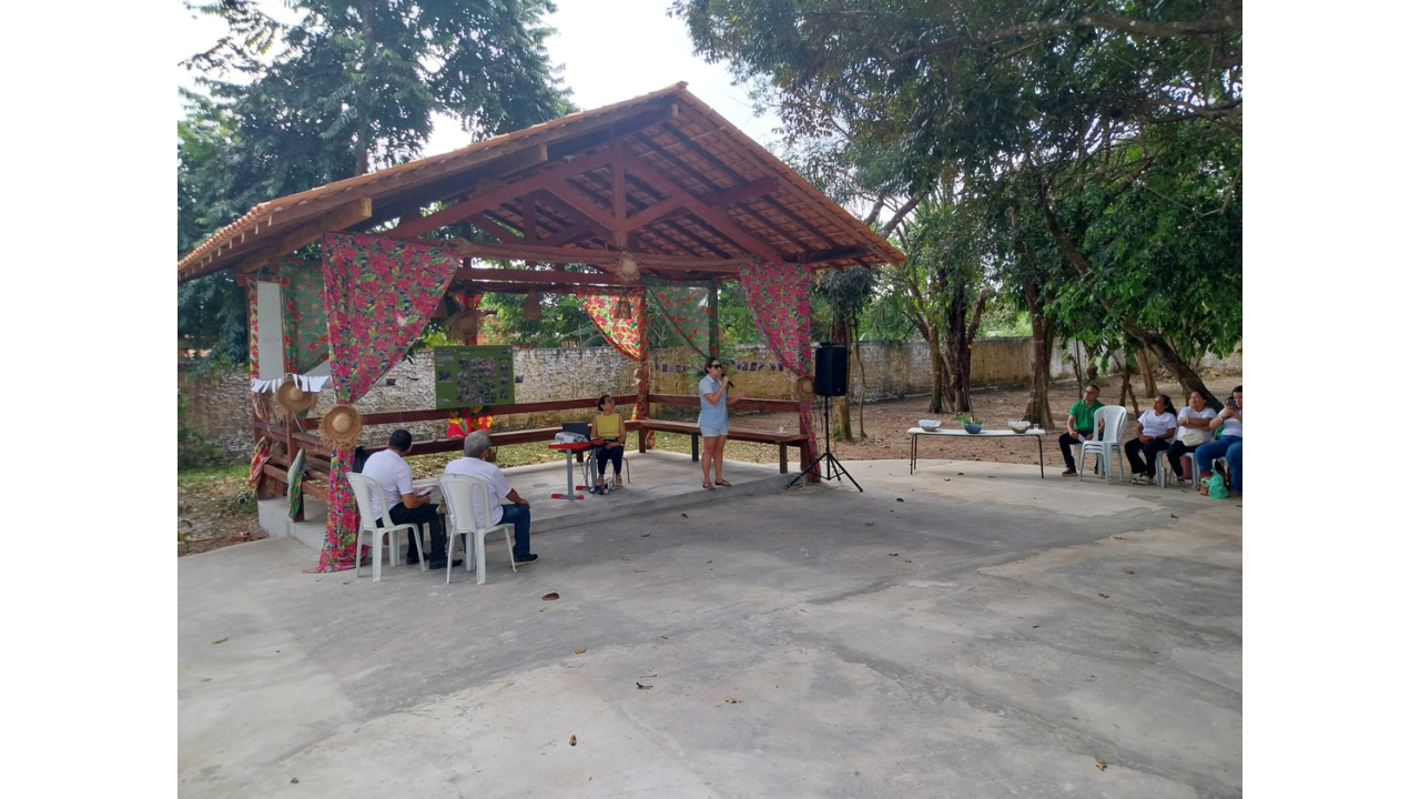 Espaço externo arborizado com uma mulher falando ao microfone diante de um grupo sentado. A apresentação ocorre sob uma estrutura de madeira decorada com tecidos florais, onde também há painel, caixa de som e mesa. Ao redor, aparecem árvores, muro e cadeiras. O chão é de concreto na cor cinza claro.