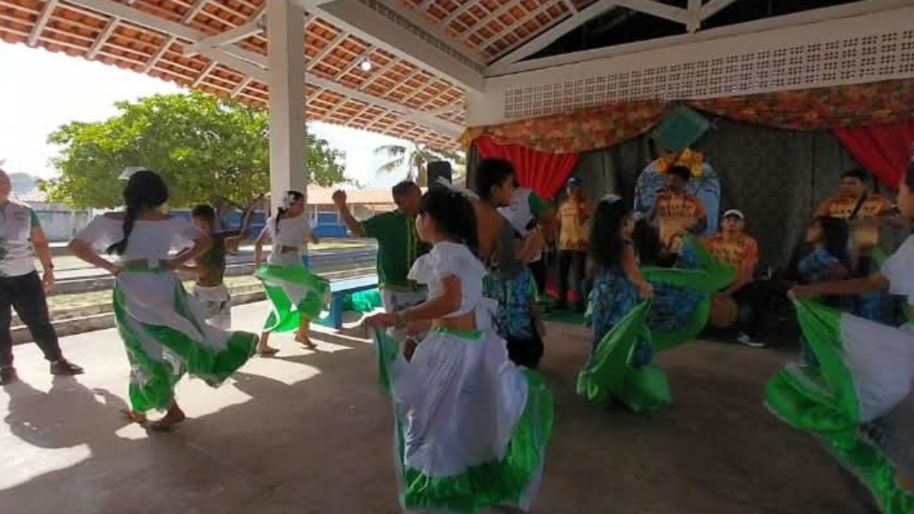 Área coberta com grupo de mulheres realizando apresentação de dança. Elas usam camisetas brancas e saias também brancas com aplicação de tecidos verdes. Elas fazem movimentos circulares, com as saias rodando a seu redor. Ao fundo, há um tecido verde com cortinas floridas e no tom de laranja, onde é possível avistar alguns músicos: um homem segura um violão e outro um microfone. O espaço possui palco, cobertura de telhas e chão de cimento.