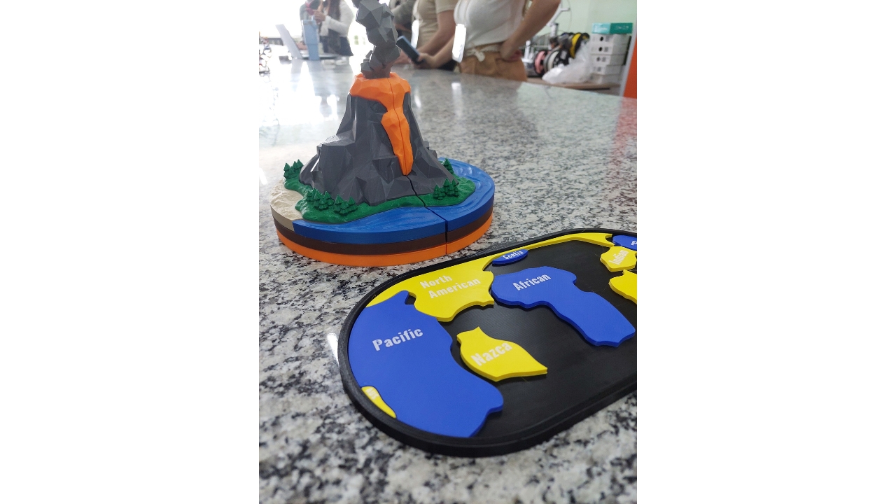 Mesa de granito de exposição em ambiente escolar. Em primeiro plano, há peças didáticas tridimensionais: um modelo de vulcão com lava laranja e um mapa nas cores azul e amarela, com fundo preto, com nomes de continentes e oceanos em inglês. As peças são, aparentemente, de plástico. Ao fundo, pessoas em pé aparecem parcialmente desfocadas.