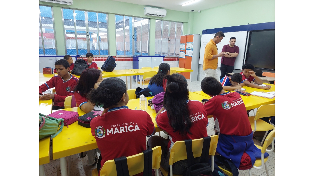 Sala de aula com estudantes sentados em mesas e cadeiras amarelas e dois adultos em pé à frente. Os alunos vestem roupas vermelhas e azuis, com a inscrição "Prefeitura de Maricá" nas costas e o brasão da cidade, enquanto os adultos conversam entre si próximos a um quadro branco e prato na frente da sala. Ao fundo, há janelas amplas e armários coloridos. As paredes da sala são da cor verde claro.