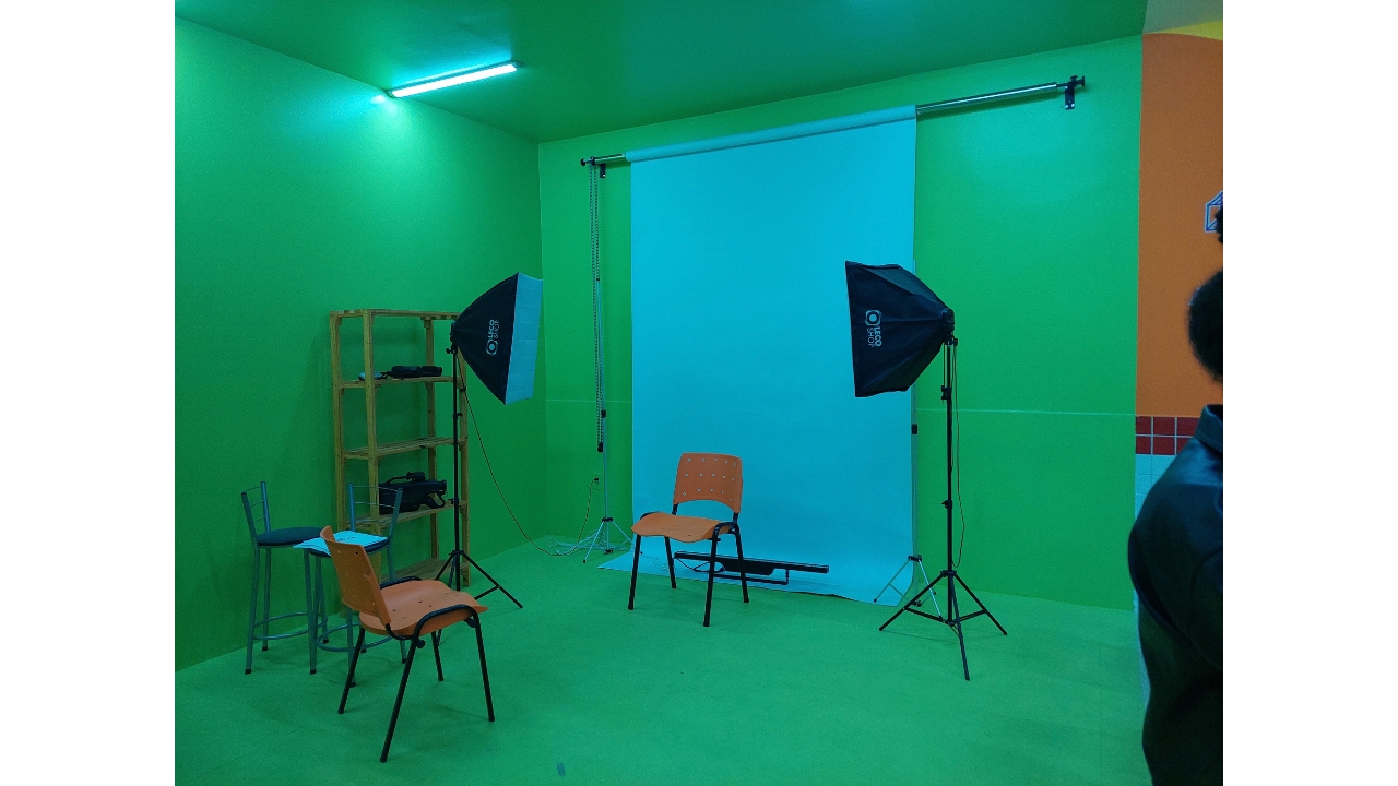 Sala verde preparada para registro fotográfico. Ao centro, há um fundo fotográfico branco, duas luzes de estúdio sobre tripés e duas cadeiras laranjas posicionadas, uma de costas para o fundo branco, centralizada, e outra de frente para a outra cadeira. À esquerda, observa-se uma estante de madeira com equipamentos.
