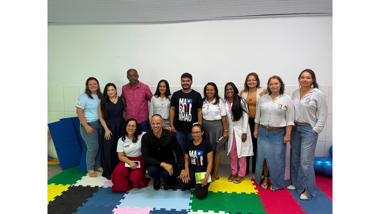 Grupo multirracial de cerca de treze adultos posa para uma fotografia sobre um piso de placas coloridas de EVA. Algumas pessoas estão em pé e outras agachadas, usando roupas casuais. Ao fundo, há uma parede branca e materiais recreativos, como colchonetes e bambolês.